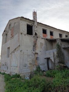Fotograf&iacute;a lateral del Molino Sarrampedra con muros agrietados, vegetaci&oacute;n invasora y se&ntilde;ales de expolio reciente en huecos y carpinter&iacute;as desaparecidas.